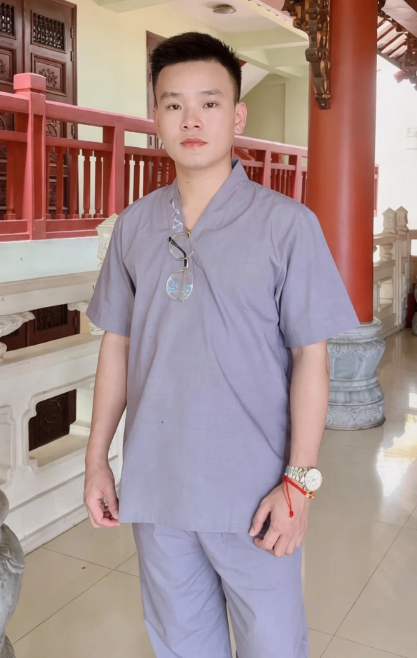 Pháp Phục Young man in light gray scrubs stands in a temple like hall with red wooden railings behind him posing for a photo