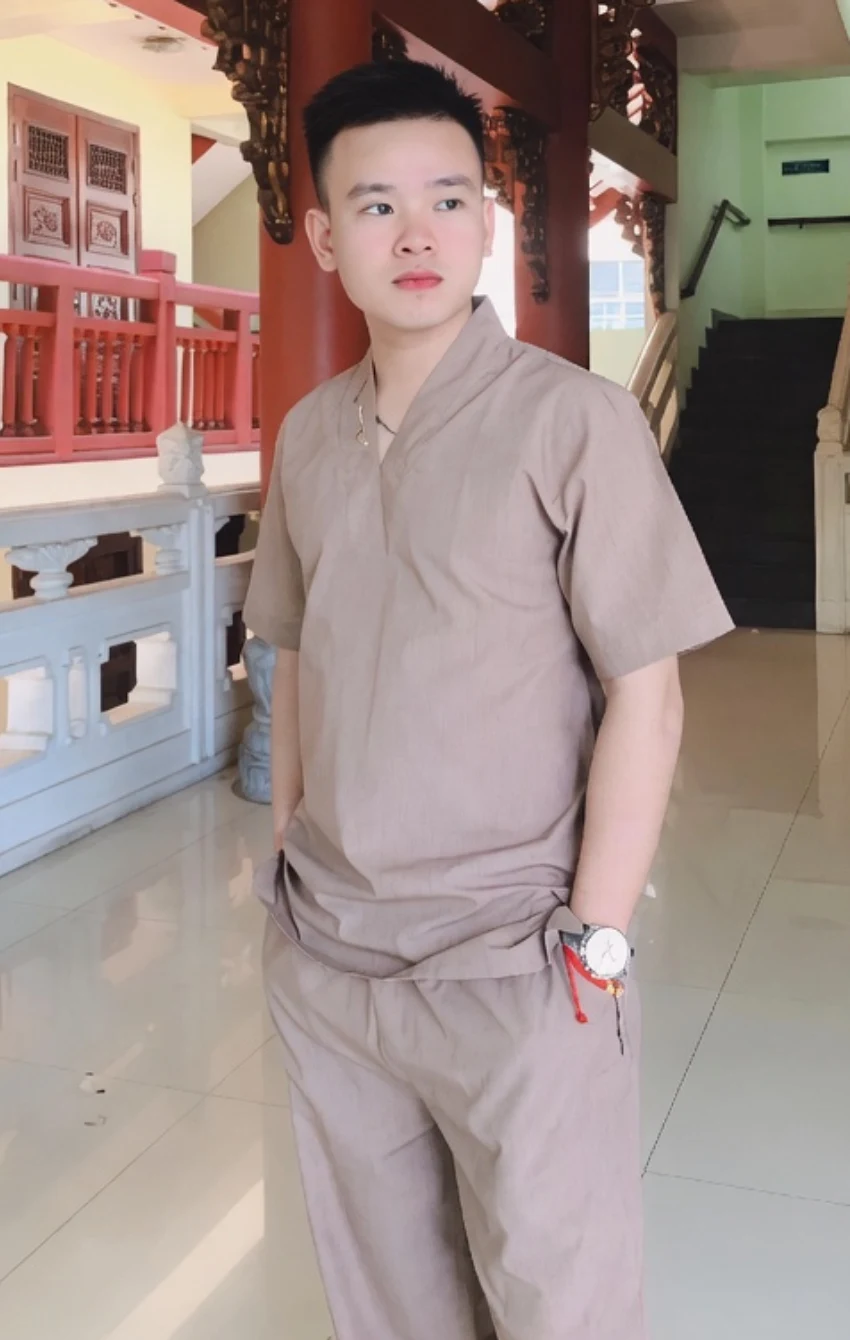 Pháp Phục Young man in a beige outfit standing in a temple like hall with red pillars and ornate woodwork