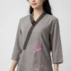 Woman wearing a light gray traditional top with dark trim, pink floral embroidery on the lower front, and three-quarter sleeves.