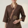 Asian woman wearing a brown wrap-style top with golden flower embroidery and a tied waist, standing against a light backdrop.