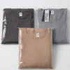 Three folded shirts in plastic packaging on a white surface, in black, gray, and tan colors with tags attached.