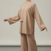Woman wearing a beige, loose-fitting tunic and wide-leg pants, standing with arms extended in a neutral studio background.