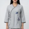 Portrait of a woman in a light gray wrap-style blouse with brown piping, side tie, and blue floral embroidery on the right side.