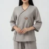 Woman wearing a gray wrap-front tunic with brown piping and embroidered rose, paired with matching gray pants.