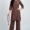 Woman modeling a brown tunic with floral embroidery on the left chest and matching wide-leg pants, wearing beige heels and holding a brown beaded necklace.