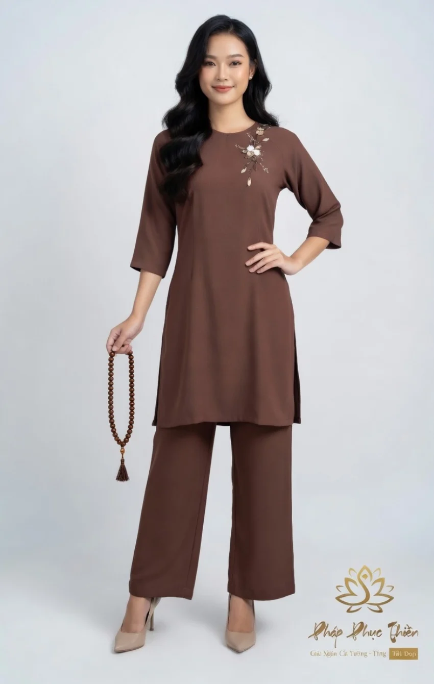 Woman modeling a brown tunic with floral embroidery on the left chest and matching wide-leg pants, wearing beige heels and holding a brown beaded necklace.