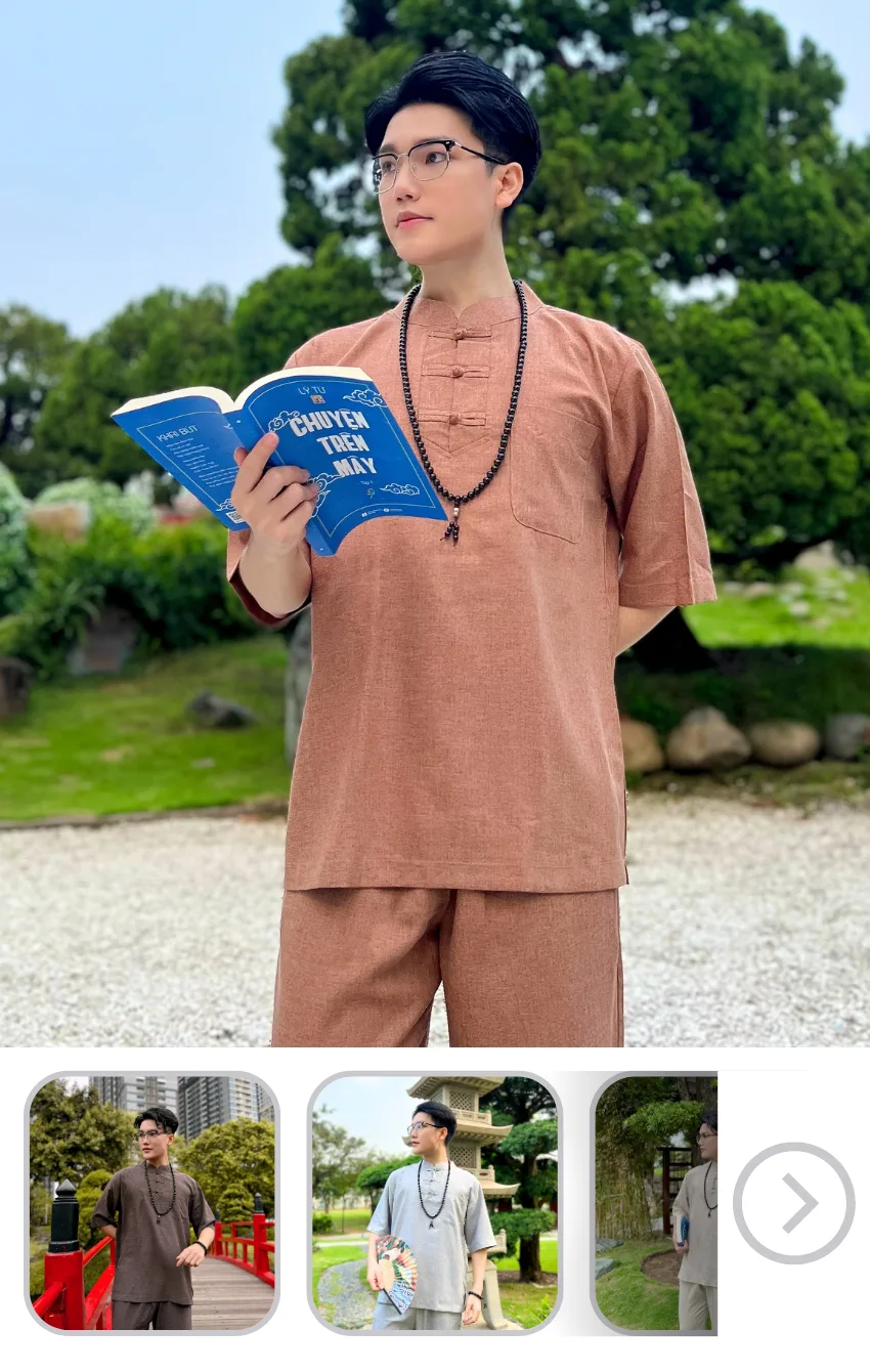 Person standing outdoors in brown traditional outfit, reading a blue book with glasses and a beaded necklace.
