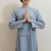 Woman in a light blue tunic and matching pants stands against a neutral wall, hands pressed together in front of her chest in a prayer-like gesture, smiling softly.