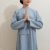 Woman in a light blue traditional outfit standing indoors with palms pressed together in front of her chest in a prayer gesture.