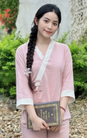 Pháp Phục Young woman in a pink traditional outfit with a long braided hair poses outdoors holding a book with gold lettering