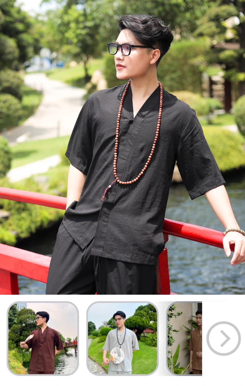 Man in black traditional-style outfit with long bead necklace leaning on a red railing beside a pond, outdoors on a sunny day, wearing sunglasses.