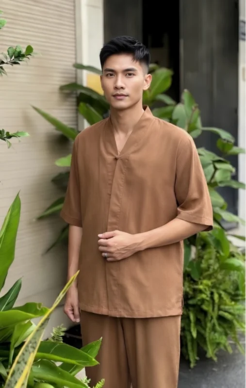 Pháp Phục Man wearing a loose brown top and matching wide leg pants stands outdoors beside a doorway with green tropical plants in the background