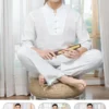 Pháp Phục Asian man in a white traditional outfit sits crosslegged on a woven cushion holding a wooden scroll in a bright room with sheer curtains