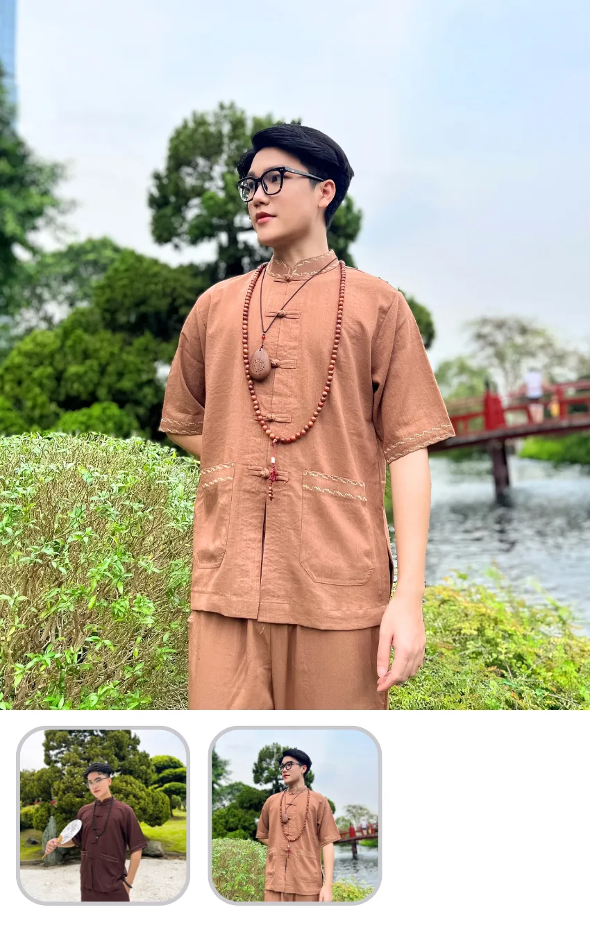 Man in brown traditional attire with glasses and beaded necklace standing by a pond with a red bridge in the background.