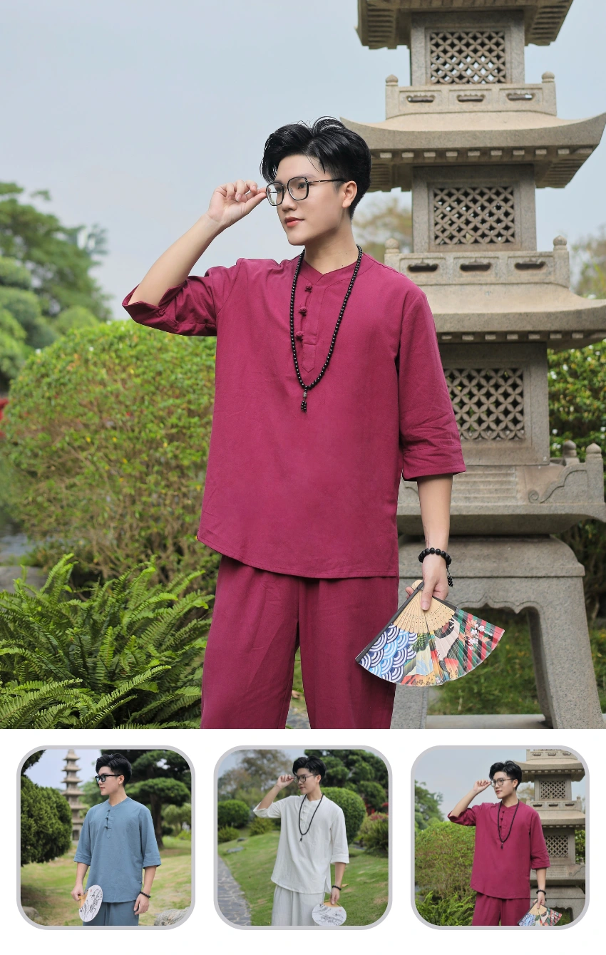 Person in maroon traditional outfit adjusts glasses while holding a patterned folding fan, standing beside a stone pagoda in a garden.