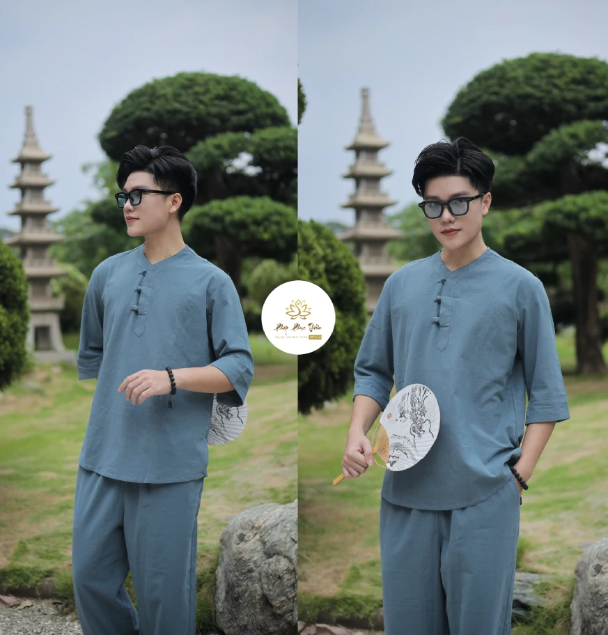 Pháp Phục Man in blue traditional attire and glasses stands in a landscaped garden with a stone pagoda in the background holding a white fan on his left side