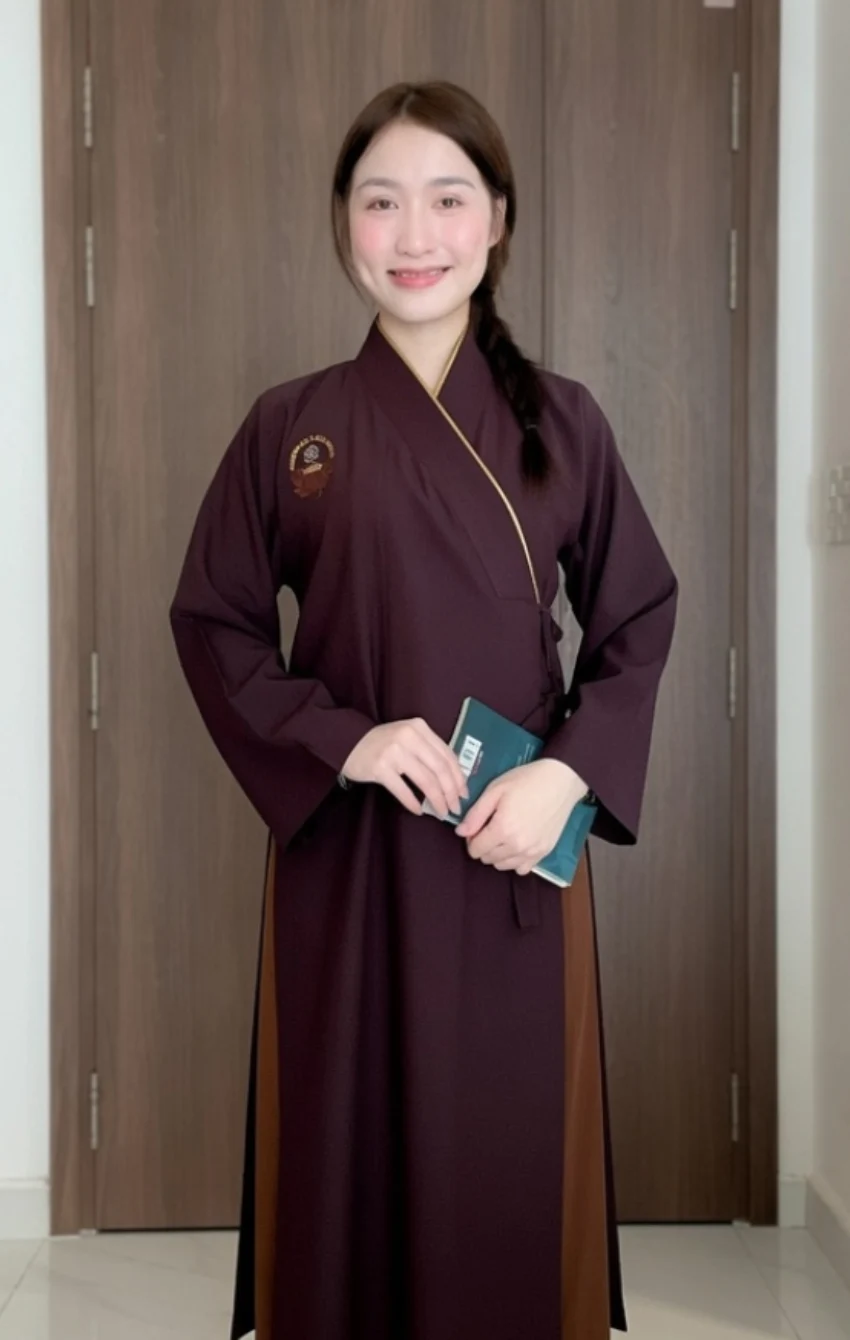 Pháp Phục Young woman in a burgundy traditional robe standing indoors in front of a wooden door holding a teal passportbooklet and smiling