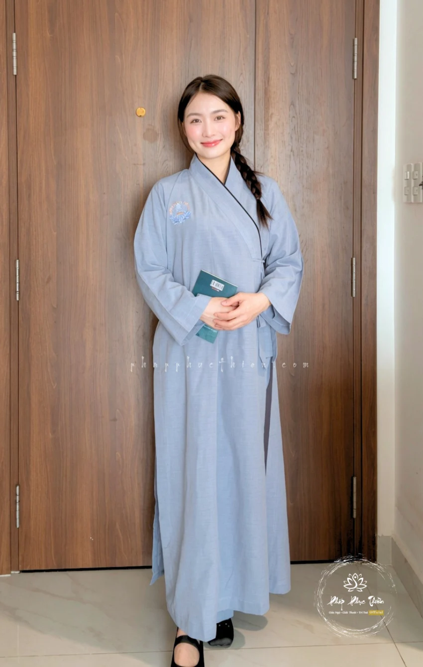 Pháp Phục Woman in a light blue kimono style robe standing against wooden doors holding a small teal book and smiling