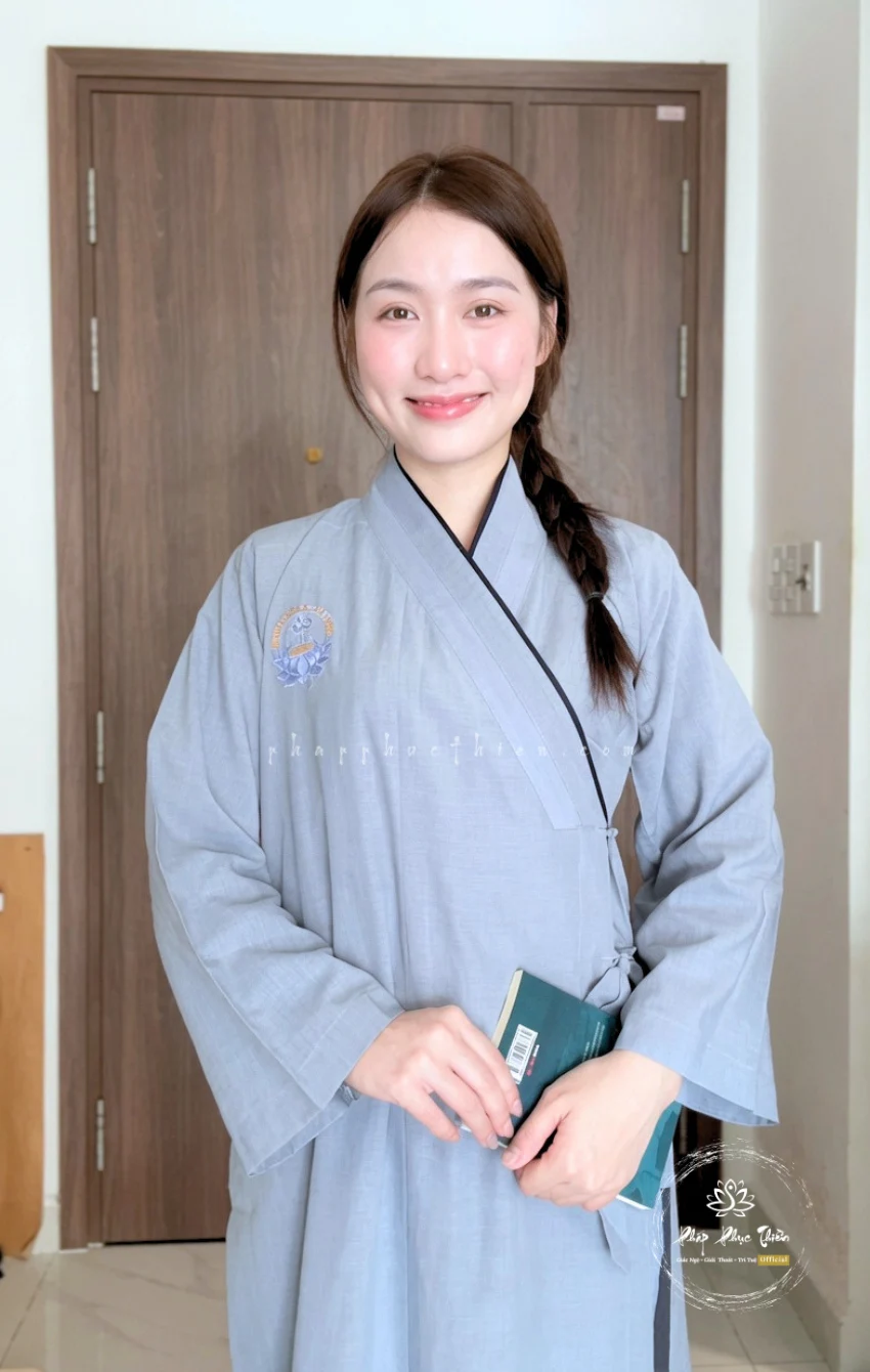 Pháp Phục Smiling woman in a light blue robe standing indoors holding a green book