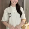 Smiling woman indoors wearing a white top with Chinese knot closures, holding a woven straw bag.