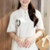Smiling Asian woman wearing a white Chinese-style tunic with a circular left-chest embroidery, holding a straw handbag indoors.