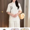 Smiling woman indoors wears a white traditional-style top and matching pants, holding a straw handbag with a bow. Emblem on the chest of the top is visible.