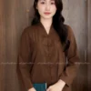 Young woman with long dark hair in a brown traditional Chinese outfit with frog closures, holding a teal book in front of wooden doors.