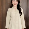 Smiling woman in a light beige traditional tunic with frog closures, standing indoors against wooden doors.