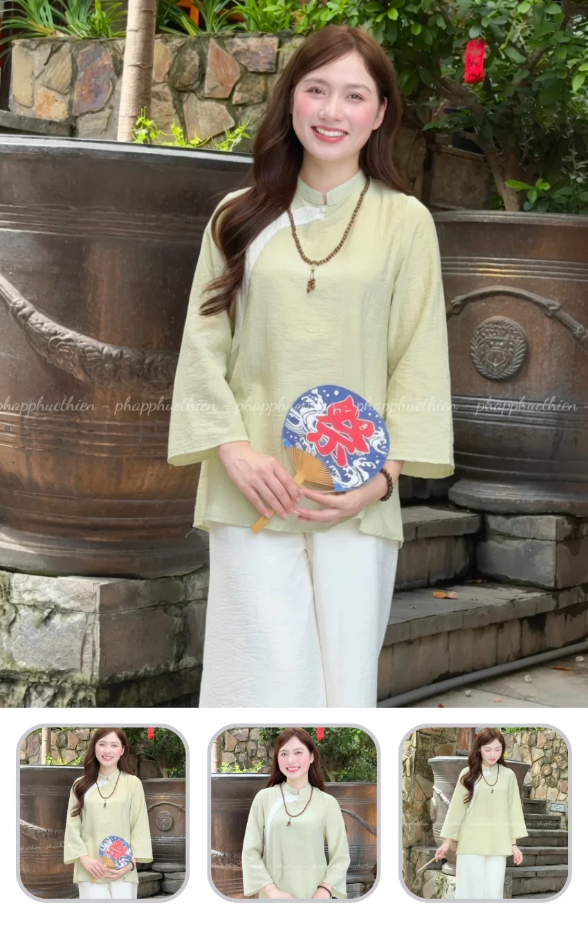 phap-phuc-dep-cho-nu Smiling woman in a pale green traditional top holds a blue decorative fan with a red symbol outdoors by a stone wall.
