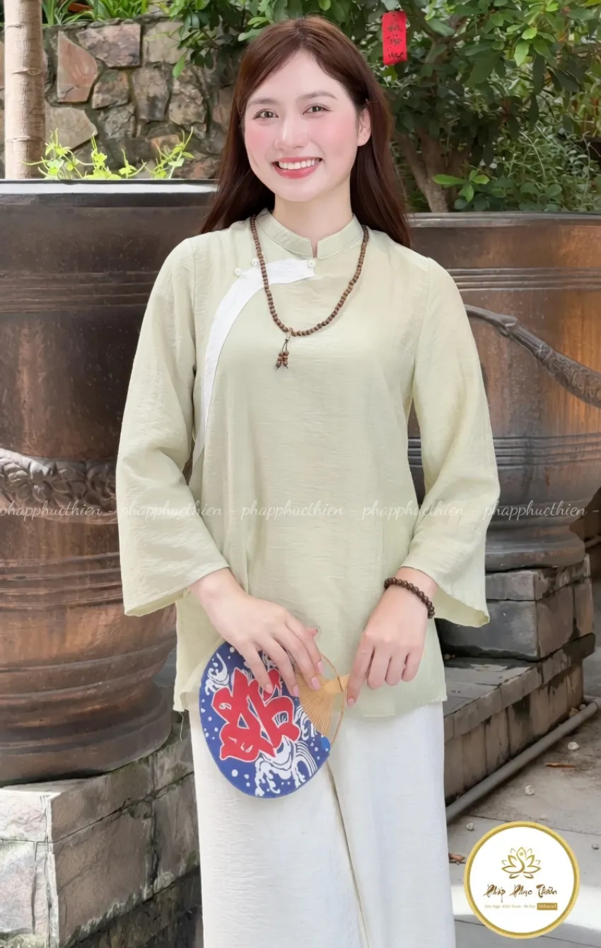 Pháp Phục Smiling woman in a pale green traditional tunic holds a round decorative fan outdoors with plants and stone wall in the background