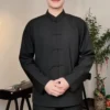 Person standing indoors in a black traditional Chinese outfit, hands gently clasped in front, neutral smile.
