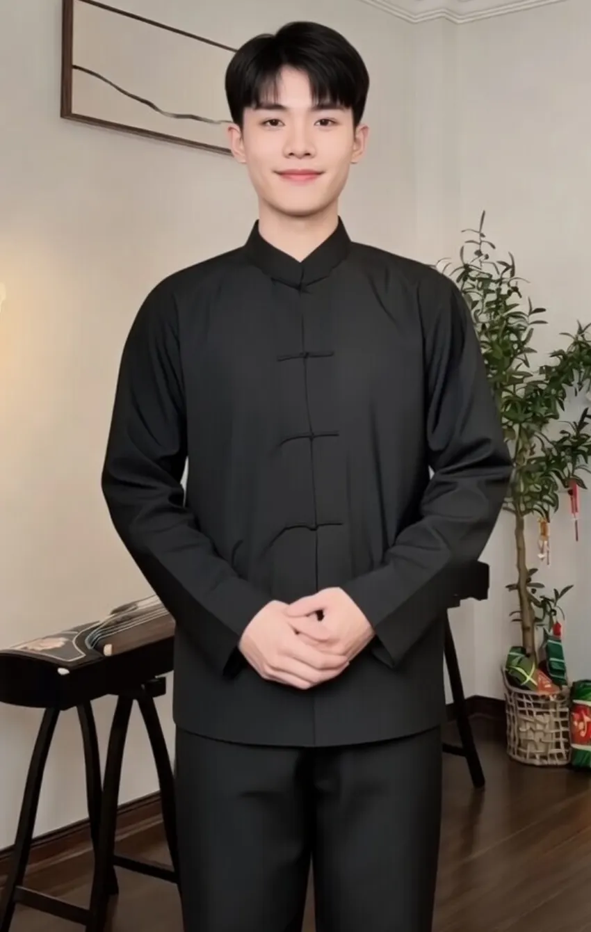 Person standing indoors in a black traditional Chinese outfit, hands gently clasped in front, neutral smile.