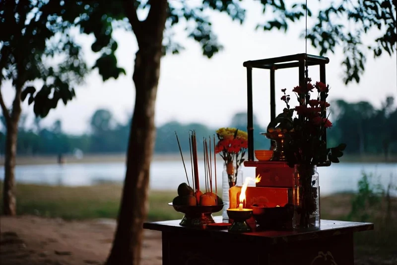 Pháp Phục Outdoor shrine by a lakeside at dusk with a lit flame incense sticks fruit offerings and flowers on a wooden table