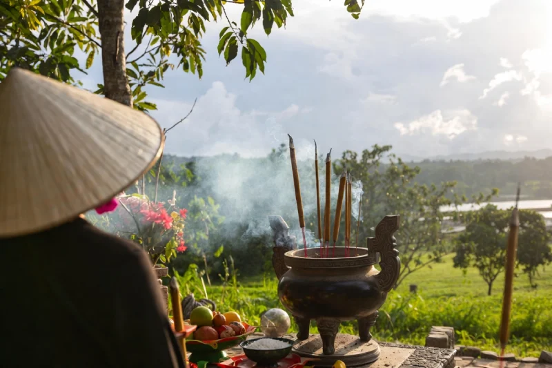 Pháp Phục Person in a conical hat at an outdoor offering ritual incense burns in a bronze censer with fruit and flowers countryside in the background