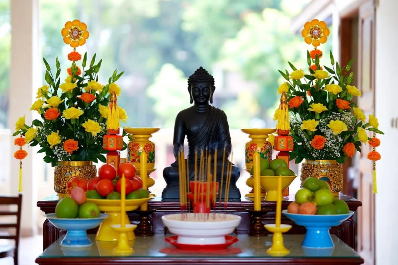 Pháp Phục Buddha statue centered on an altar with fruit bowls candles incense and vibrant yellow and orange flower arrangements on either side
